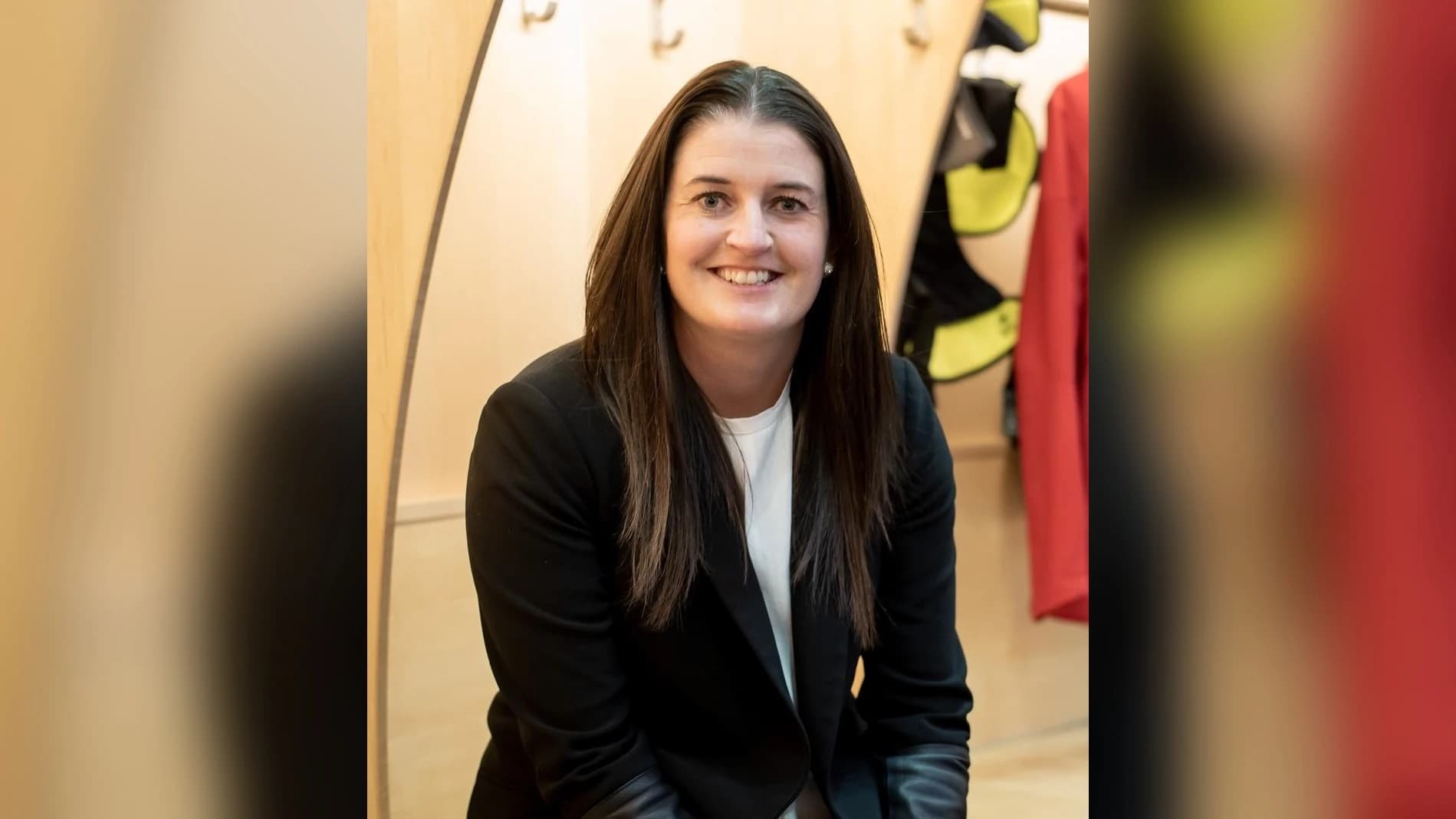 Gina Kingsbury, neue General Managerin beim ÖEHV, lächelt in die Kamera. Sie trägt einen schwarzen Blazer über einem weißen Shirt und hat lange, dunkle Haare. Im Hintergrund ist eine helle Holzkabine mit Garderobenhaken und aufgehängter Ausrüstung zu sehen.