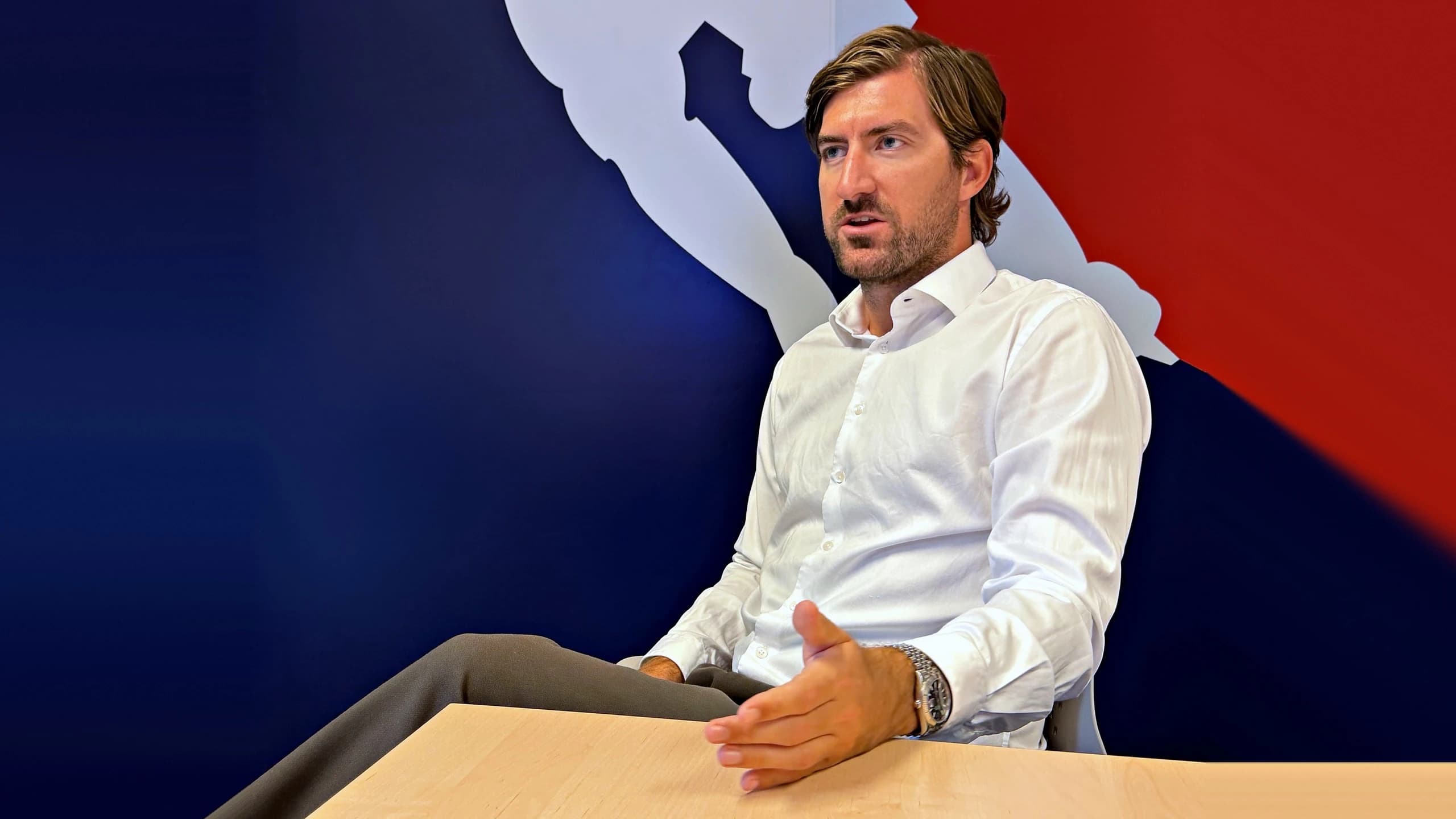 ÖEHV-Geschäftsführer Alexander Helwig sitzt im weißen Hemd an einem Holztisch und gestikuliert im Gespräch mit einer Hand. Im Hintergrund ist eine blau-rote Wand mit der weißen Silhouette eines Eishockeyspielers zu sehen.