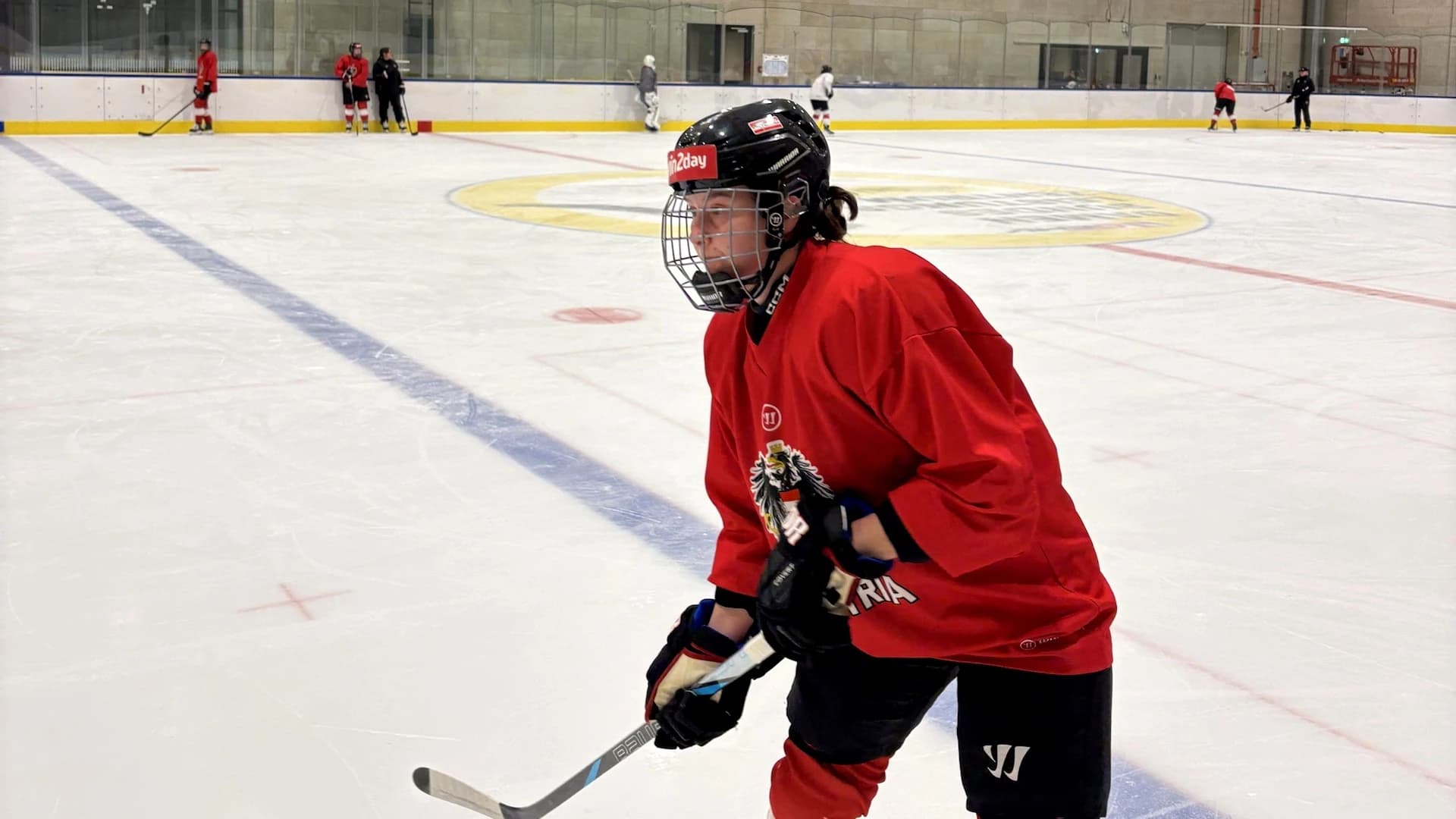 Eishockeyspielerin des Frauen-Nationalteams in rotem Trainingstrikot mit Helm und Gitter beim Training; im Hintergrund weitere Spielerinnen auf dem Eis.