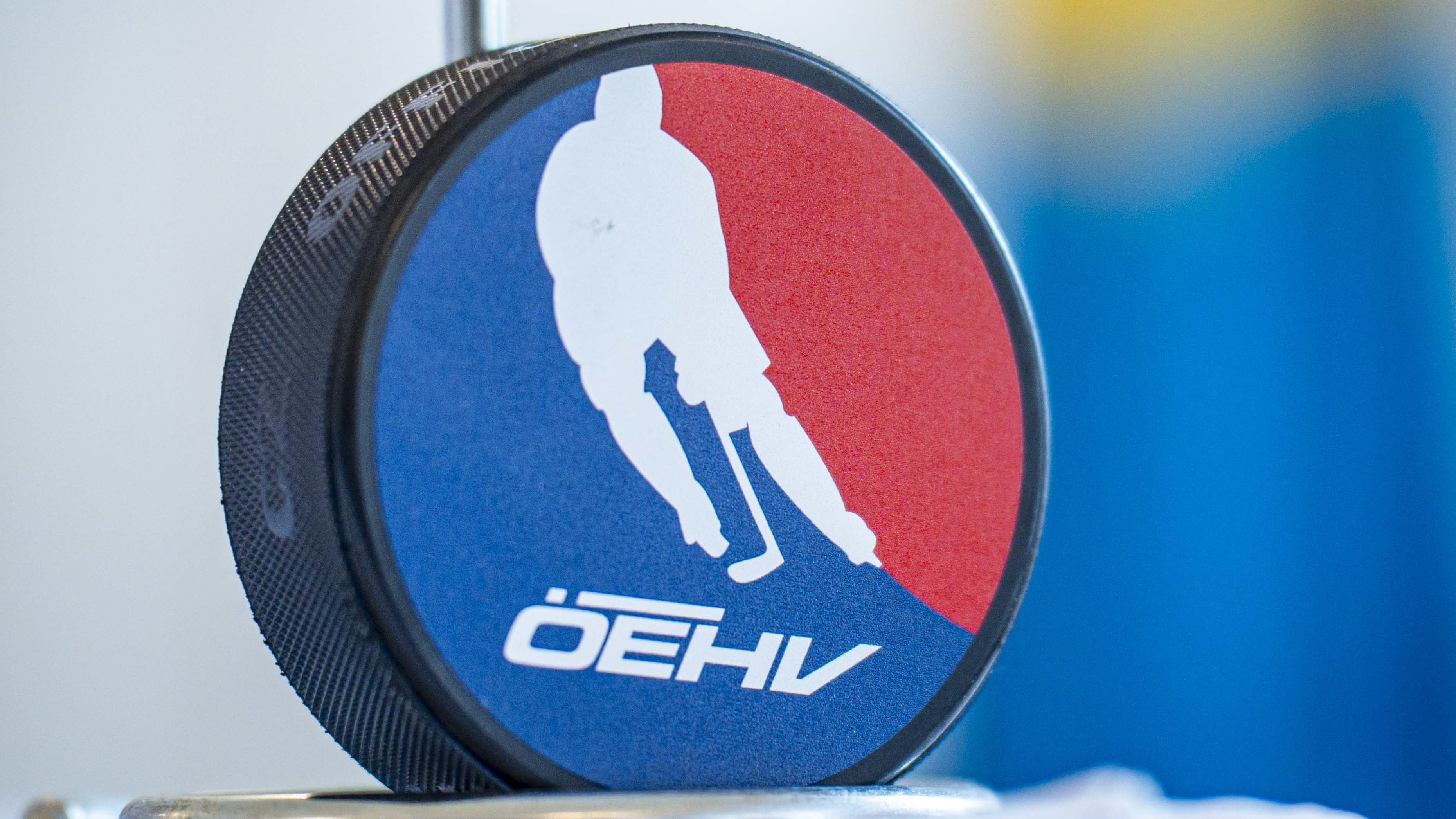 Nahaufnahme eines Eishockeypucks mit ÖEHV-Logo: Die Seitenfläche zeigt einen stilisierten Eishockeyspieler in Rot und Blau sowie den Schriftzug ÖEHV. Der Puck steht auf einer reflektierenden Fläche vor unscharfem, farbig verschwommenem Hintergrund.