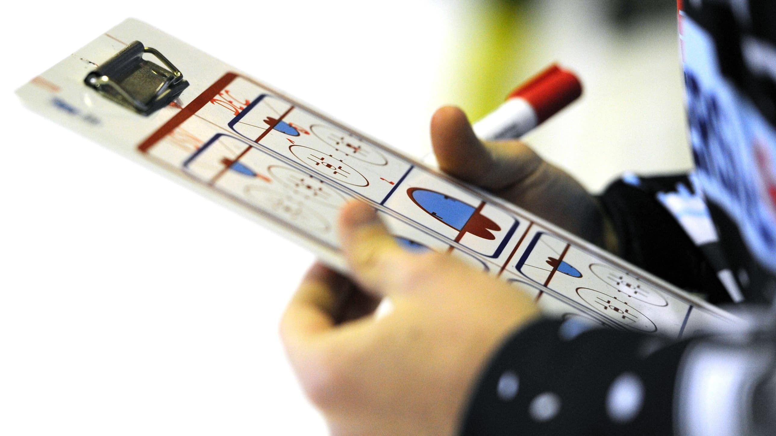 Nahaufnahme einer Hand, die eine Taktiktafel für Eishockey hält. Auf der weißen Tafel sind mehrere Eishockeyfelder abgebildet, in der anderen Hand ein roter Whiteboard-Stift.