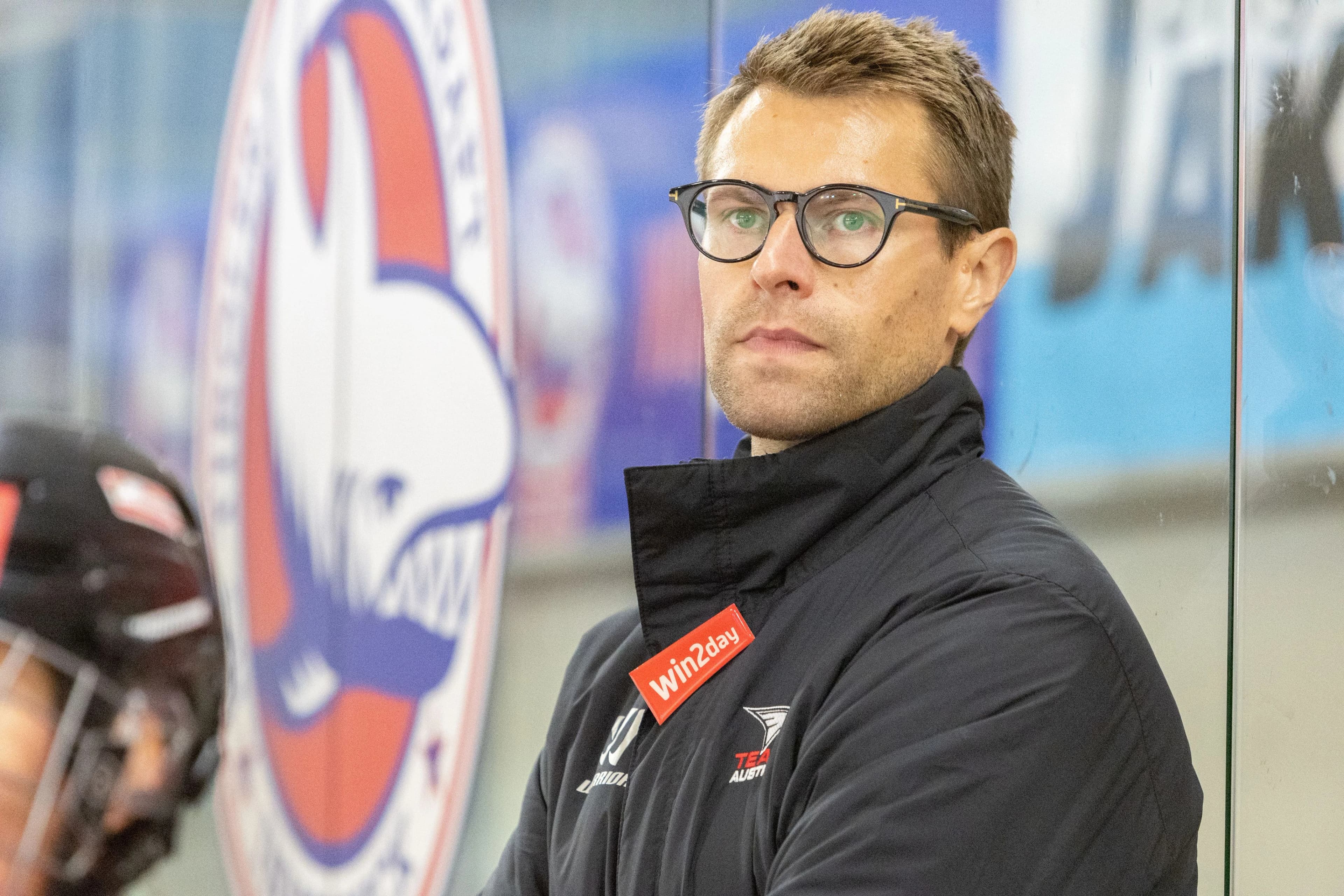 Ein Mann mit Brille und dunkler Jacke mit roten Details steht vor einer Logowand und blickt konzentriert zur Seite.