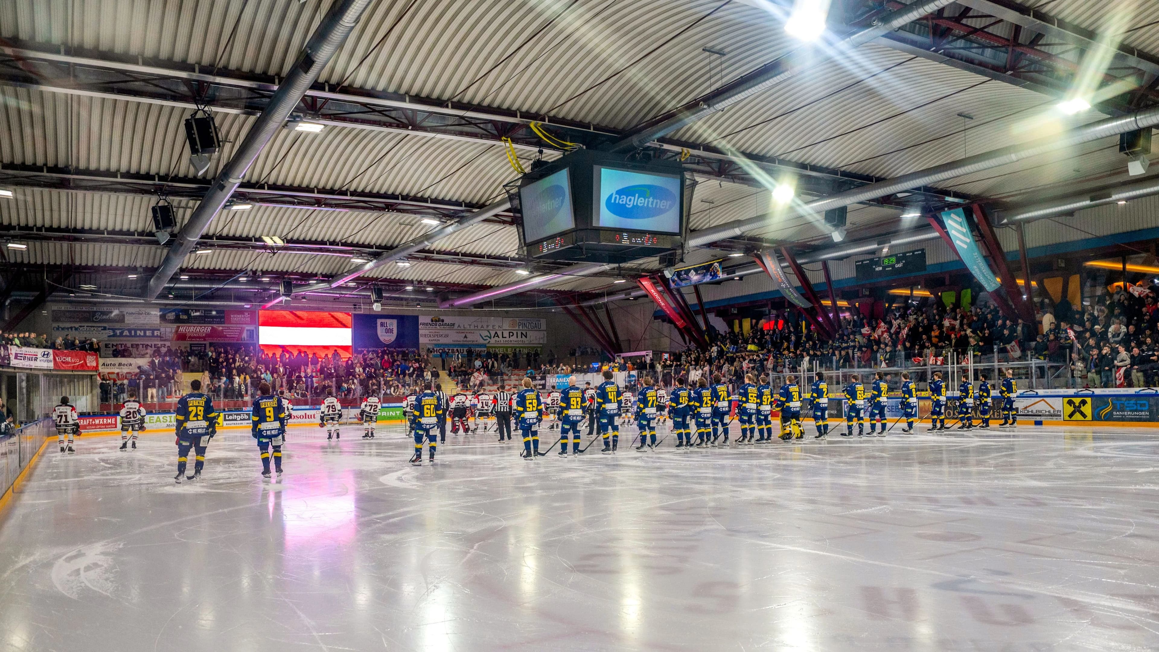 Eishockeymannschaften in blau-gelben und weiß-roten Trikots stehen zur Nationalhymne aufgestellt am Spielfeldrand einer Eishalle, im Hintergrund gefüllte Zuschauertribünen und eine Anzeigetafel mit Sponsorenlogo.