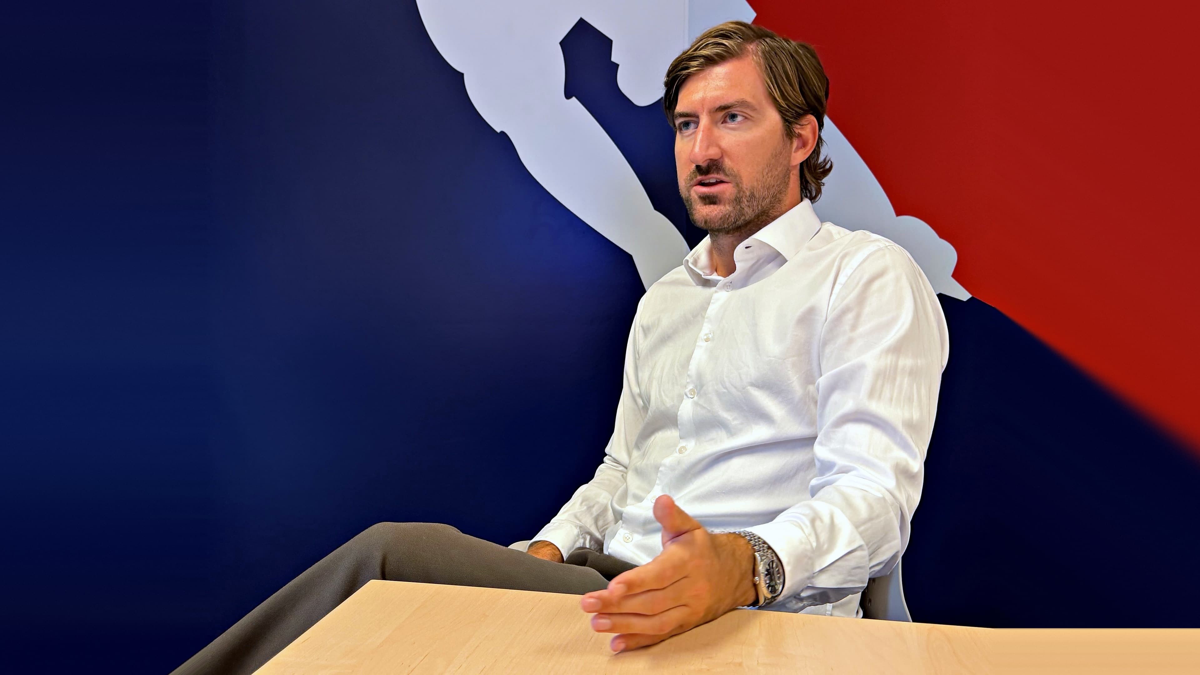 ÖEHV-Geschäftsführer Alexander Helwig sitzt im weißen Hemd an einem Holztisch und gestikuliert im Gespräch mit einer Hand. Im Hintergrund ist eine blau-rote Wand mit der weißen Silhouette eines Eishockeyspielers zu sehen.