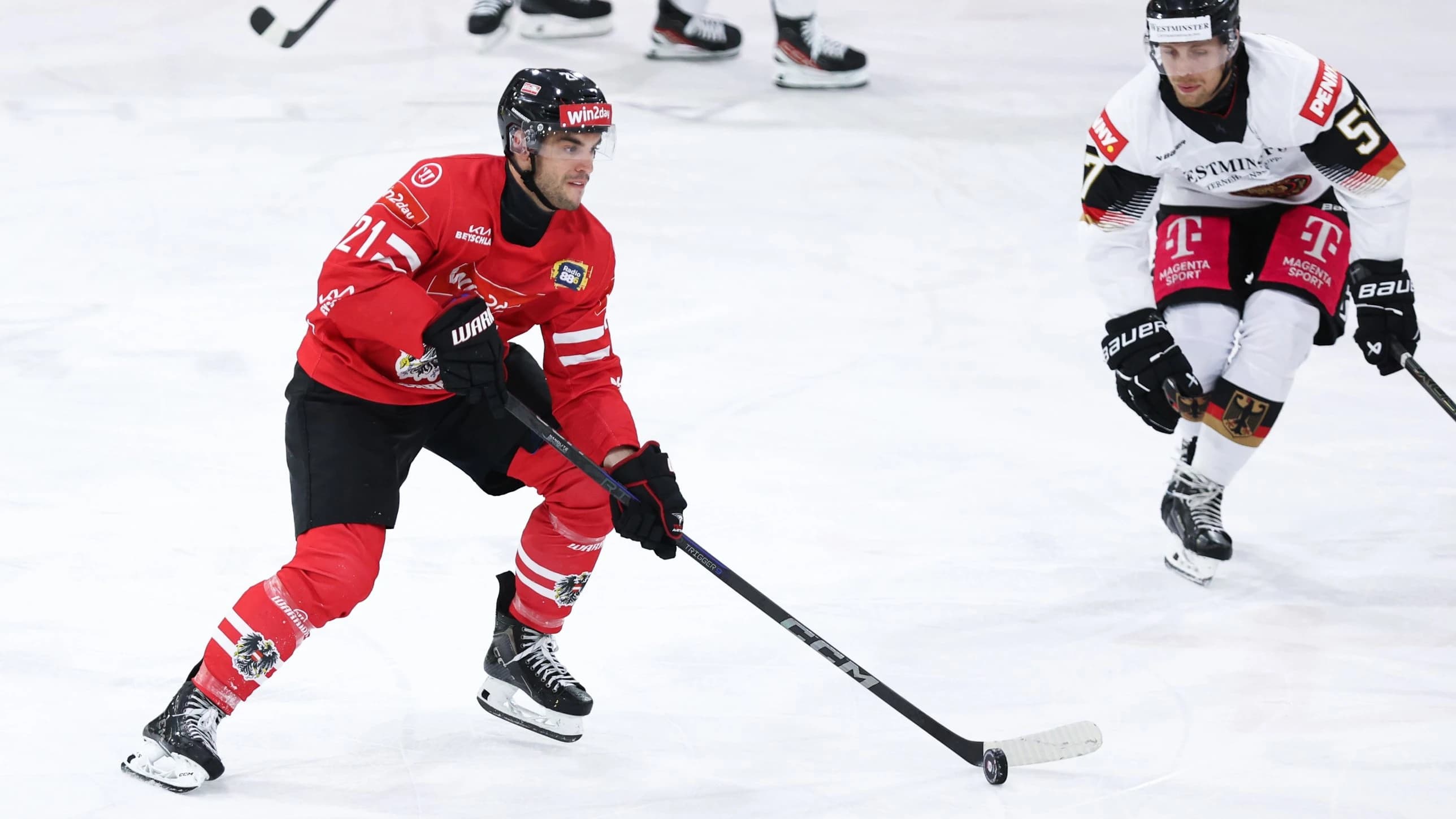 Ein Spieler des österreichischen Männer-Nationalteams in rotem Trikot mit der Nummer 21 führt den Puck mit dem Schläger über das Eis und blickt nach vorne. Rechts im Bild verfolgt ihn ein deutscher Spieler in weißem Trikot mit schwarz-roten Elementen. Im unscharfen Hintergrund sind die Beine weiterer Spieler zu sehen.