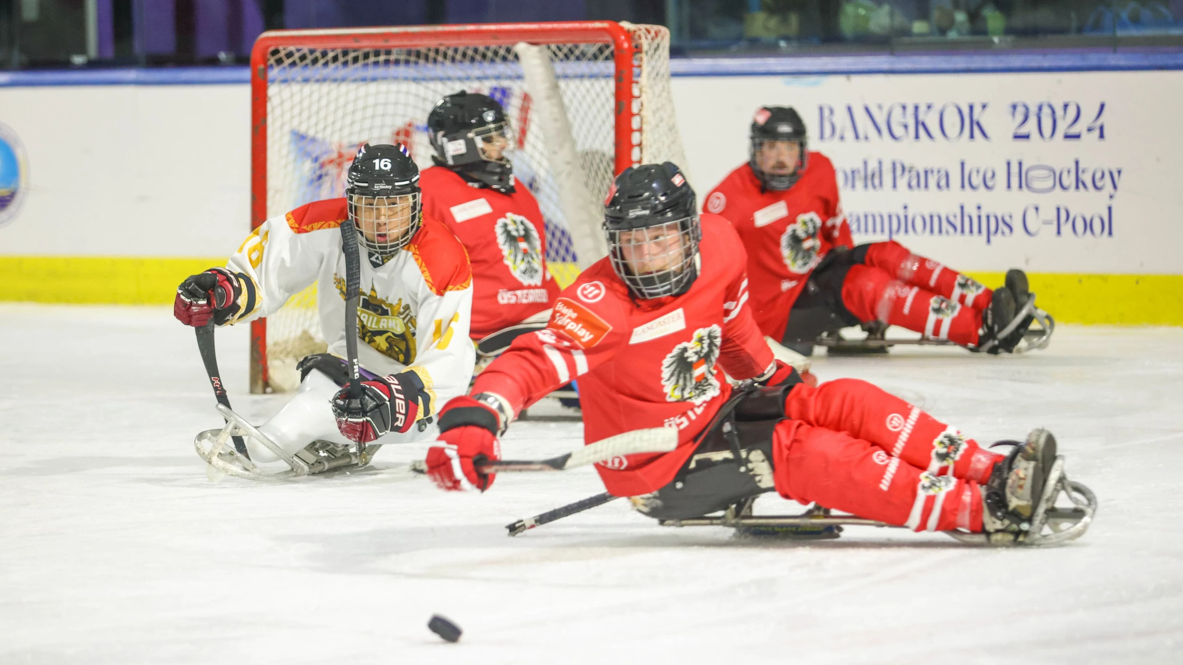 Österreichischer Para-Eishockey-Spieler im roten Trikot kämpft im Sledge um den Puck gegen einen Spieler aus Thailand, im Hintergrund weitere Spieler vor dem Tor, Bande mit Aufschrift 'Bangkok 2024 World Para Ice Hockey Championships C-Pool'.