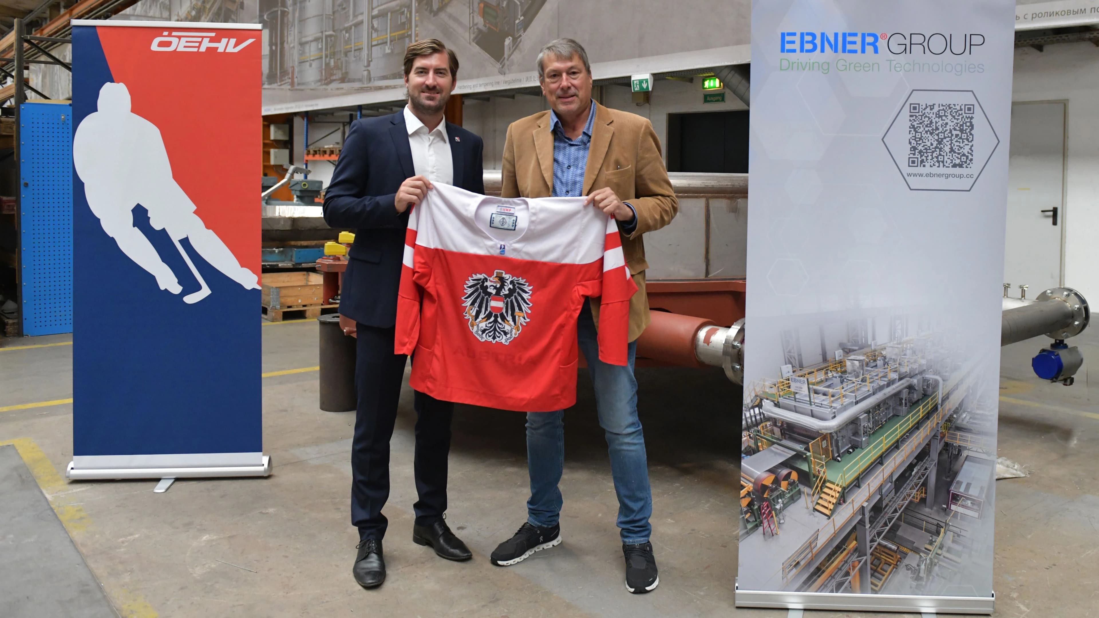 Zwei Männer präsentieren gemeinsam ein rot-weißes Damen-Nationalteam-Trikot Österreichs mit Doppeladler-Wappen in einer Industriehalle, flankiert von Roll-ups des ÖEHV und der EBNER GROUP.