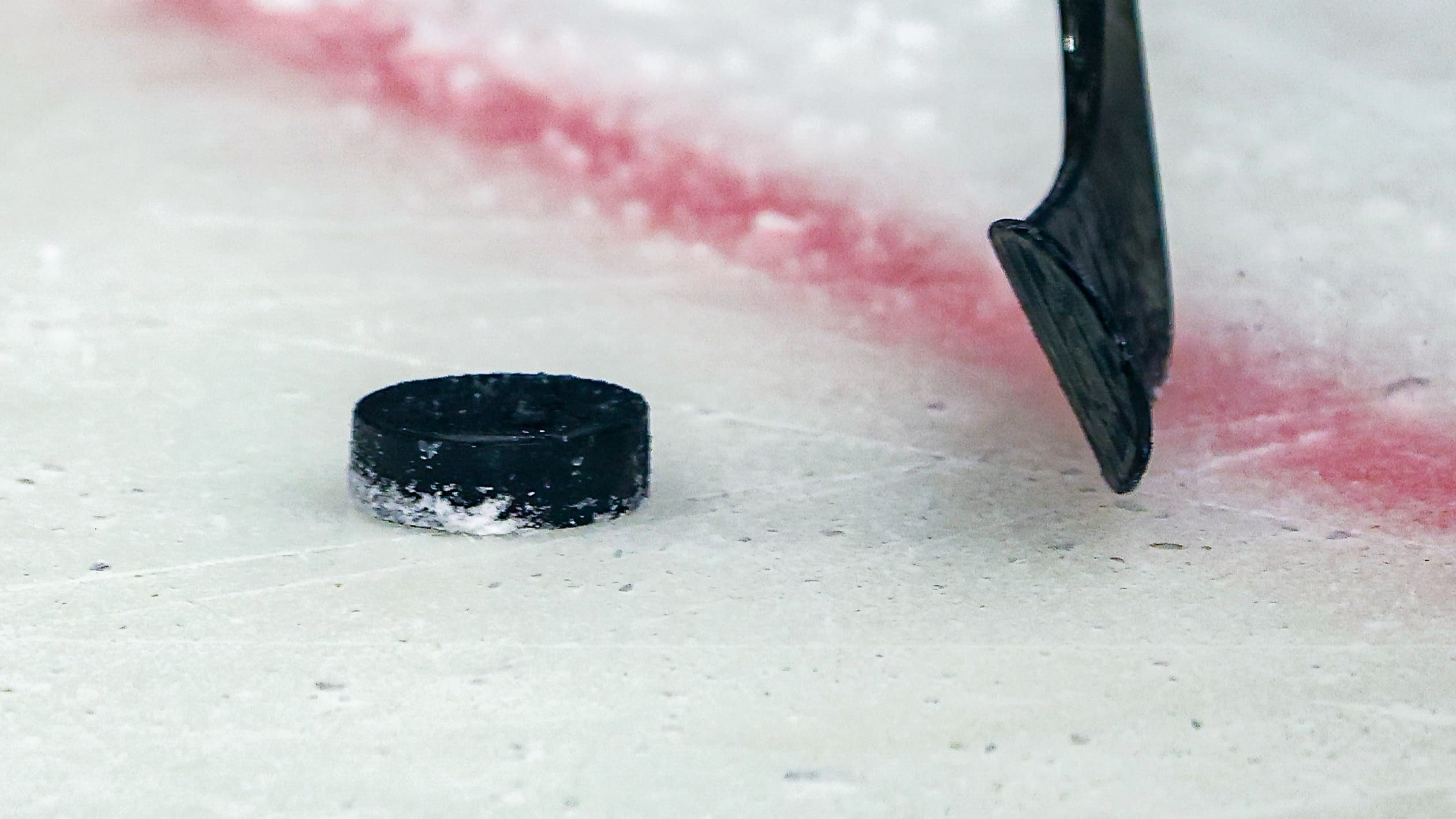 Ein schwarzer Puck liegt auf dem Eis neben einer roten Linie, rechts daneben schwebt die Kelle eines Eishockeyschlägers knapp über der Eisfläche.