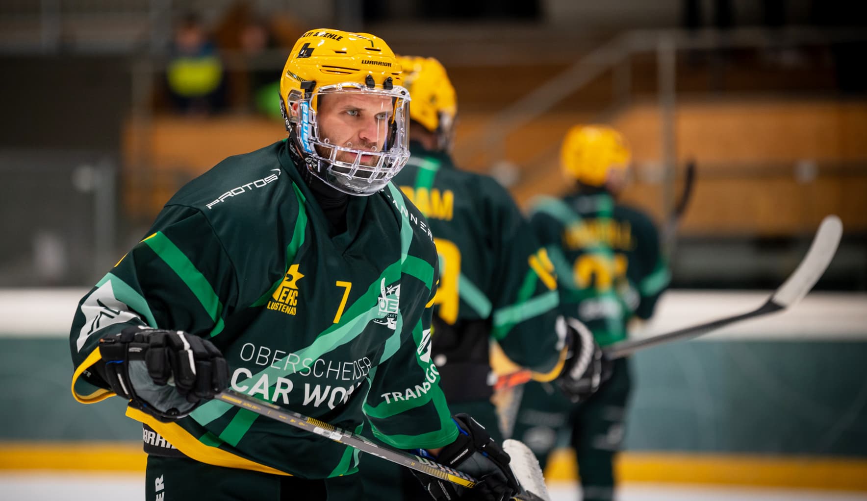 Ein Spieler des EHC Lustenau mit der Nummer 7 steht in dunkelgrünem Trikot und gelbem Helm mit Vollvisier konzentriert auf dem Eis. Im unscharfen Hintergrund sind zwei Teamkollegen zu sehen.