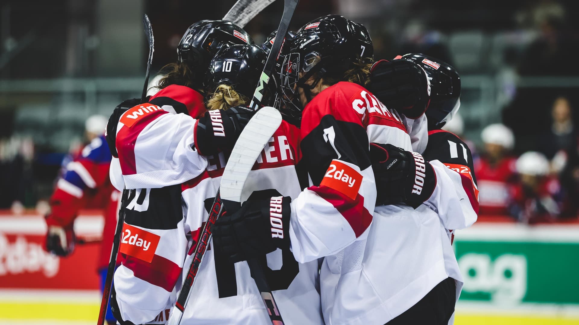 Spielerinnen des österreichischen Frauen-Nationalteams in weißen Trikots mit roten und schwarzen Elementen jubeln eng umarmt in einer Traube auf dem Eis, die Helme aneinandergelehnt und die Schläger in die Höhe gestreckt. Im unscharfen Hintergrund sind die Spielerinnen der gegnerischen Mannschaft in roten Trikots zu sehen.